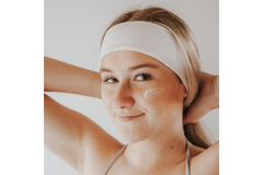 woman wearing headband with face cream on cheek holds her hair and looks at camera